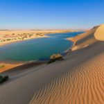 A serene panoramic view of The Magic Lake in Fayoum Oasis, showing its shimmering blue waters surrounded by golden desert dunes under a clear sky
