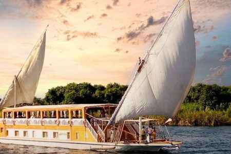 An elegant, traditional Dahabiya sailboat with large, white canvas sails gently gliding on the Nile River