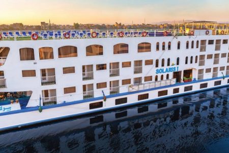 Un grand bateau de croisière sur le Nil blanc et bleu, nommé "SOLARIS I" sur sa proue, naviguant sur une étendue d'eau calme. Le bateau a plusieurs ponts avec de nombreuses fenêtres, des balcons et une rangée de chaises longues bleues sur le pont supérieur.