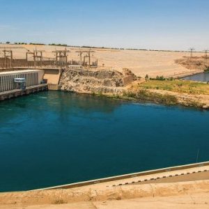 Vue panoramique du Haut Barrage d'Assouan, montrant le lac artificiel du barrage en premier plan avec ses eaux bleues profondes. À gauche se trouve une longue structure en béton du complexe hydroélectrique, reliée à des pylônes électriques. En arrière-plan, la vaste structure en terre du barrage s'étend sous un ciel bleu clair.